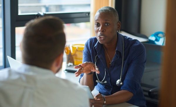 Patient and doctor talking in person in an office room with the back of the patients head visible and the in full view, speaking assertively and gently to the patient.